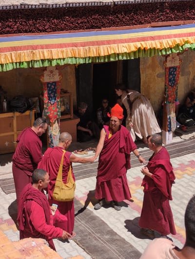 In Pibiting, India, a group of Buddhist monks and visitors are gathered outside a building adorned with colorful, striped awnings. Several monks in crimson robes are the central figures. One monk, wearing an orange conical hat and glasses, stands facing forward, holding a hand out. Another monk, with a yellow bag slung over his shoulder, is shaking hands with a third monk who is also wearing a crimson robe. In the background, a woman in a light-colored, long dress is seen entering a doorway, and other individuals are visible in the shadows. The ground is paved with a mosaic of colored tiles and striped rugs. The building features decorative pillars and a textured, dark roofline.