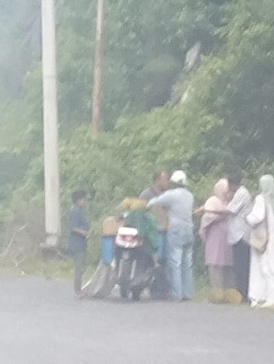 In Pasi, Indonesia, a group of people are gathered around a motorcycle. The group includes several men and women, some of whom are wearing traditional Islamic attire. They appear to be engaged in conversation or a transaction, with some individuals touching the motorcycle. The environment is rural, with lush greenery and trees in the background, suggesting a roadside location. The lighting is overcast, indicating a cloudy day.
