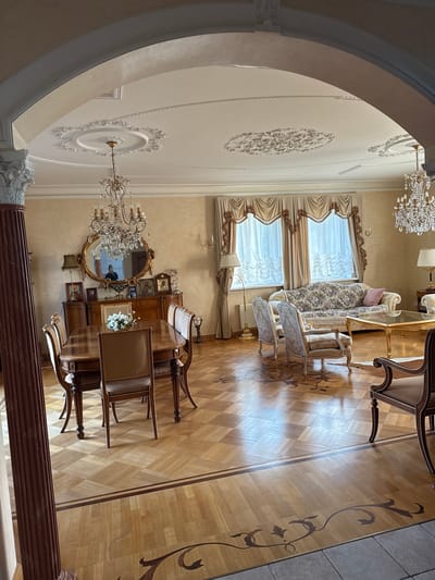 The image shows an opulent dining and living area in a home in Yukki, Russia. The room features ornate chandeliers, plush furniture, and intricate ceiling moldings. A large wooden dining table with six chairs is in the foreground, set for a meal. In the background, a floral-patterned sofa and two armchairs are arranged around a glass coffee table. The room is decorated with religious icons and a large ornate mirror above a sideboard. The flooring is a polished wood parquet with decorative inlays. The overall impression is one of wealth and traditional elegance.