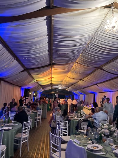 This media file depicts a formal indoor event, likely a wedding reception or a banquet, held within a draped tent. The interior is adorned with white drapes that create a flowing, textured ceiling, illuminated by strings of small lights and dramatic blue and orange lighting effects. Chandeliers are visible, adding to the celebratory atmosphere.
The scene features several round tables set with green tablecloths, white chairs, and floral arrangements, indicating a dining event. People are seated at the tables and some are standing and mingling, suggesting a social gathering. The attendees appear to be dressed in smart casual to formal attire. The lighting suggests the event is taking place in the evening or at night. The overall mood seems festive and elegant. No visible text is present in the image. The location context provided is Baliovtsi, Bulgaria.