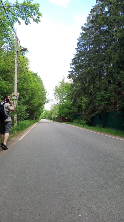 The image depicts a road in the city of Дубки, Russia. The road stretches into the distance, bordered by greenery on both sides. On the left, there's a man walking and talking on his cell phone. He's wearing a patterned shirt, shorts, a backpack, and black shoes. He stands next to a utility pole with a streetlamp and a sign. Lush green trees and bushes line the left side of the road. On the right, tall evergreen trees are present behind a green fence. The sky is mostly bright and white.