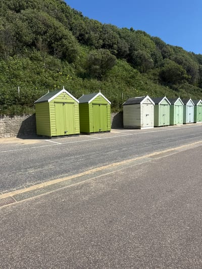The image shows a row of beach huts along a promenade in Bournemouth, United Kingdom. The huts are painted in various shades of green and white, with traditional pitched roofs. They are situated on a paved area next to a road and a grassy slope covered in dense foliage. The sky is clear and blue, indicating a sunny day. The scene is quiet and appears to be unoccupied. The huts are lined up neatly, suggesting they are part of a rental or private collection. The overall impression is one of a picturesque coastal setting.