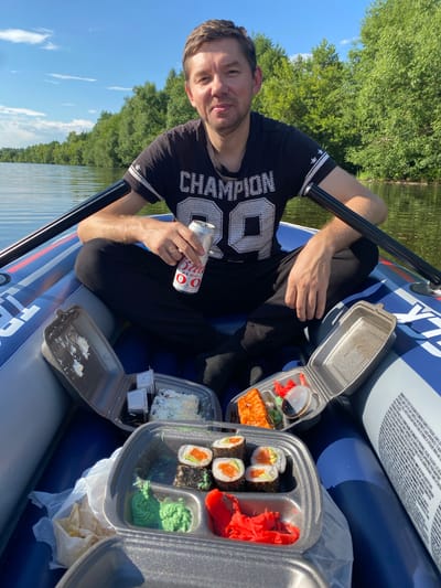 A man is in an inflatable boat on a body of water, possibly a lake or river, surrounded by trees. He is wearing a black t-shirt with white lettering and stripes on the sleeves, black pants, and has a ring on his left hand. He is holding a can of Bud Zero, which has "ZERO" in red letters and "BUD" in white letters against a red background. In front of him, there are two open takeaway containers of sushi. One container has several pieces of sushi rolls, some with salmon and avocado, and pickled ginger. The other container has a piece of salmon and some rice. There are small white containers for soy sauce. The sun is shining, indicating it is daytime, and the sky is blue with some clouds. The man is looking at the camera with a slight smile. The text on the boat reads "TR", and some warning text is visible on the inflatable. The location is Chulki-Sokolovo, Russia.