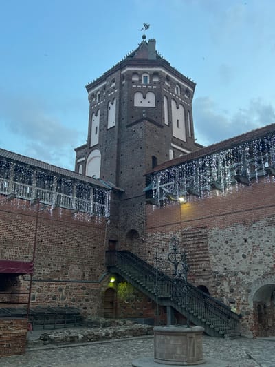 The image displays an interior courtyard view of the Mir Castle Complex in Мірскі сельскі Савет, Belarus, under an overcast blue sky. Dominating the scene is a tall, octagonal tower constructed of dark red brick with contrasting lighter arched and rectangular window recesses, crowned by a red-tiled roof and a dark weathervane. An aircraft is faintly visible in the upper left sky. Surrounding structures include brick walls with elevated wooden walkways, festooned with numerous strands of illuminated white icicle-style string lights. The courtyard floor is paved with irregular cobblestones. In the mid-ground, a circular stone well with a detailed, dark metal wellhead is centrally positioned. To the right, a dark metal staircase with wooden treads ascends along a brick wall to an upper doorway; a yellow light illuminates an arched doorway below the stairs. On the left, partially obscured by a dark red canopy, are low stone foundations and several low, rectangular, dark-colored covers. No individuals are present in the scene.