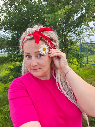 A light-skinned woman with blonde dreadlocks, wearing a red bandana and a bright pink t-shirt, is posing outdoors. She holds a white daisy with a yellow center against her right eye, with the stem extending behind her head. The woman is looking directly at the camera with a slight smile. The background consists of green foliage, trees, and a glimpse of a blue structure. The image appears to have been taken in the village of Novoe Mart'yanovo, Russia.