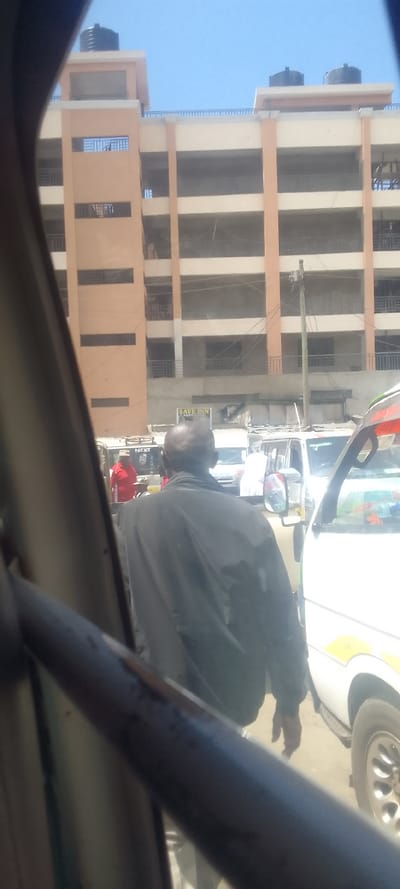 Framed from within a vehicle, the image depicts a daytime street scene in Naivasha, Kenya. A bald male, wearing a dark jacket, is centrally positioned with his back to the viewer, appearing to move past parked or slow-moving white minivans, consistent with Kenyan matatus. His right hand is partially visible. To his left, another individual in a red shirt and a light-colored hat stands near a vehicle.

The background is dominated by a multi-story building with light-toned walls, contrasting orange-brown vertical architectural elements, and multiple balcony openings secured with railings. Several black water storage tanks are visible on the building's rooftop, which also features a perimeter railing. At the building's ground level, a commercial sign for "SAVE INN CHIPS" is visible. A utility pole with multiple overhead lines stands in the mid-ground, slightly to the right of center, in front of the building. The sky above is clear and blue.