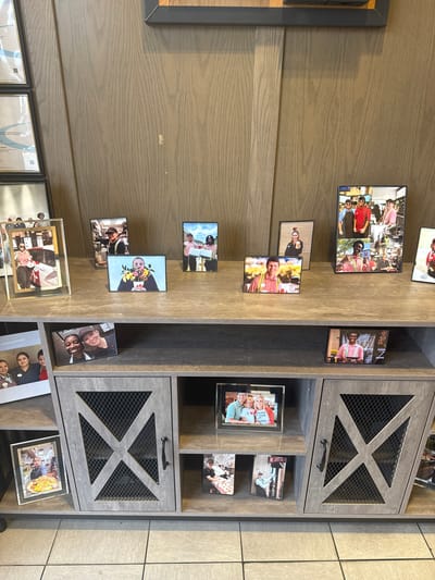 The scene shows a wooden wall decorated with framed photos and a wooden shelving unit with cabinets. The photos on the wall are a mix of personal snapshots, possibly of people associated with the location. The cabinets in the shelving unit have a mesh screen design. The background includes a glimpse of a tiled floor. The image appears to be taken in an interior space in Stone Mountain, United States of America.