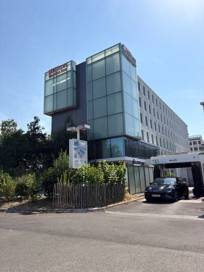 The image shows the Hilton Garden Inn hotel in Rungis, France, on a sunny day. The hotel is a multi-story building with a modern glass facade on the upper floors and a white exterior on the lower floors. A black car is entering a car wash bay located to the right of the hotel. Lush green bushes and trees surround the hotel, and a simple wooden fence lines the edge of the property. A signpost with what appears to be an advertisement or information is also visible near the entrance. The sky is clear and blue.