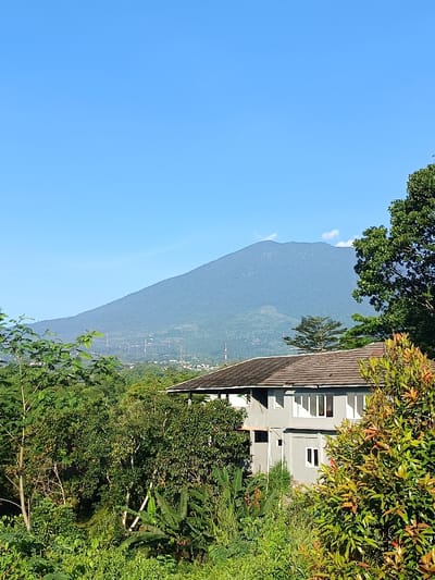 The image displays a daytime landscape in Cugenang, Indonesia. A large, green-vegetated mountain dominates the background beneath a clear blue sky. Power line towers and communication masts are visible on the mountain's lower slopes and in the distant valley, alongside scattered buildings. In the mid-ground, a two-story residential building with a brown tiled roof and light grey walls is partially visible, featuring multiple white-framed windows. The foreground is filled with dense green foliage, including various trees, shrubs, and banana plants, which partially obscure the residence. No human figures or active interactions are observed within the frame.