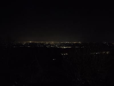 The image displays a panoramic nighttime view from an elevated position in Villa di Gemmano, Italy. The upper two-thirds of the frame consists of a dark, unlit sky. Below this, a distant urban area is visible, characterized by a horizontal band of numerous small, scattered artificial lights, predominantly yellow and white, extending across the mid-ground. A diffuse glow is perceptible above the most concentrated areas of illumination, indicative of light pollution. The immediate foreground is dark and indistinct, featuring the silhouetted forms of leafless vegetation, resembling tree branches or shrubs, visible on the lower left and right edges of the frame, partially obscuring the distant lights. No individuals, explicit actions, or direct interactions are discernible.