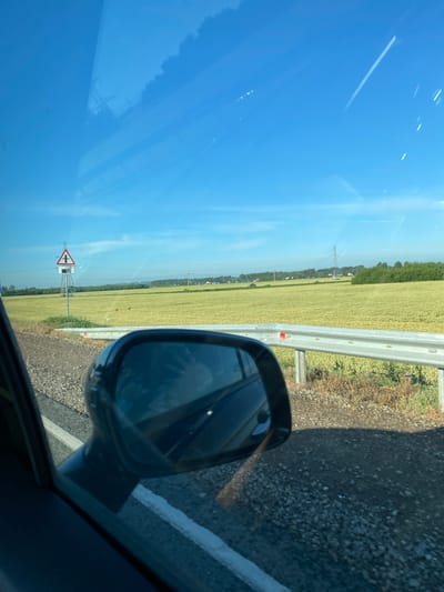 The image depicts a view from inside a vehicle, looking out the side window towards a rural landscape. A car's side-view mirror is prominently featured in the foreground, reflecting a blurry image of what appears to be a person. Outside the car, to the left, a triangular road sign indicating an intersection is visible. Beyond the road, a vast field of yellow grain stretches towards a treeline in the distance. Power lines and utility poles traverse the horizon. The sky is a clear, bright blue with a few wispy clouds. The road itself is paved with gravel and asphalt, with a white line marking the edge. The scene is set in a rural area, likely a highway or country road, in Vasilyevka, Russia, given the presence of Russian road signs and the general landscape characteristics.