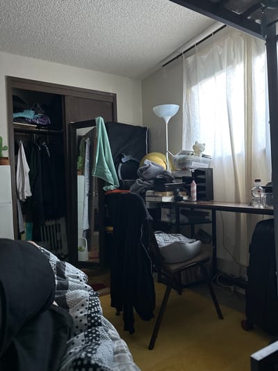 The image depicts a cluttered bedroom. A closet filled with clothes is visible on the left. In the center, a desk is piled with various items including papers, a lamp, headphones, and a pink spray can. A chair with a pillow on it sits in front of the desk. A window with sheer curtains is on the right, letting in natural light. The floor is covered with a yellow carpet. The overall impression is of a lived-in, disorganized space. The location is unknown.