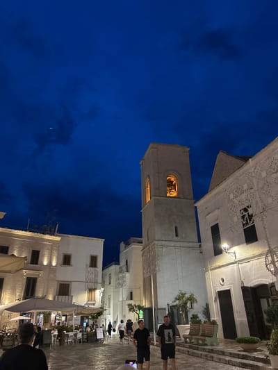The image depicts a nighttime scene in Polignano a Mare, Italy, likely in the historic center. In the foreground, several people are walking along a cobblestone street. To the left, outdoor seating for a restaurant is visible under large umbrellas, with patrons seated at tables. In the mid-ground, a tall, light-colored bell tower dominates the scene, with a warm light emanating from its arched openings, illuminating the interior bells. Adjacent to the bell tower is a white building with intricate decorative white latticework along its facade. Further to the left and right are more white buildings, illuminated by artificial lighting. The sky above is a deep blue, with some clouds and what appears to be a small airplane or drone visible high in the sky. The overall atmosphere suggests a charming European town square at dusk or early evening.