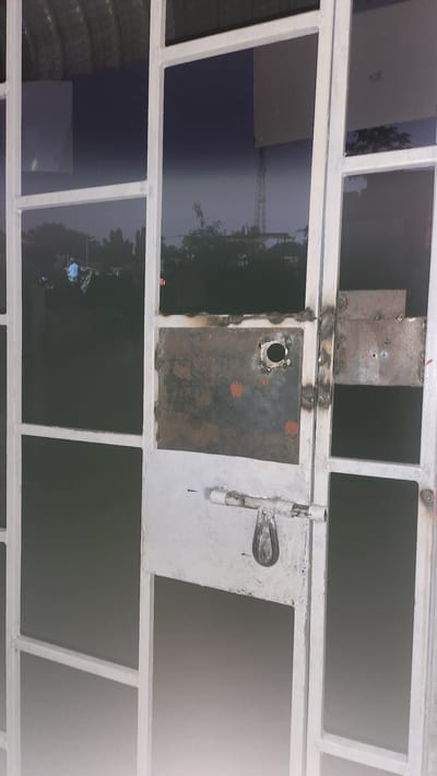 The image shows a white metal gate with several panels. The gate is in an open position, revealing a dark, possibly nighttime, environment beyond. Reflections on the gate panels show a faint image of trees and a distant tower structure. A simple metal latch mechanism is visible on one of the panels, appearing to be in the unlocked position. The gate is showing signs of wear and tear, with visible rust spots and rough welds on some of the metal plates. The overall scene suggests an outdoor location, likely a barrier to an establishment or property. The location is identified as Kipala Mpakani, Tanzania.