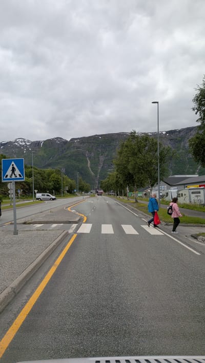 The image depicts a pedestrian crossing on a road in Mosjøen, Norway. Two people are crossing the street: an adult in a blue jacket carrying a red bag and a child in a pink jacket. They are in the middle of the road, walking from right to left relative to the viewer.
The scene is set with a paved road and a pedestrian crossing marked with white stripes. A yellow line runs along the edge of the road. To the left of the road, there is a pedestrian crossing sign with a blue background and a white silhouette of a person walking. Beyond the sign, a white van is parked.
In the background, there are trees and buildings, including what appears to be a commercial area with some trailers or caravans parked outside. Majestic, rocky mountains with patches of snow dominate the skyline, suggesting a mountainous region. The sky is overcast with clouds, indicating a cloudy or possibly rainy day. The time of day appears to be daytime, given the visibility. The overall mood seems neutral, focusing on the everyday activity of crossing the road in a scenic environment. There is no discernible text in the image, apart from the text on the pedestrian crossing sign.