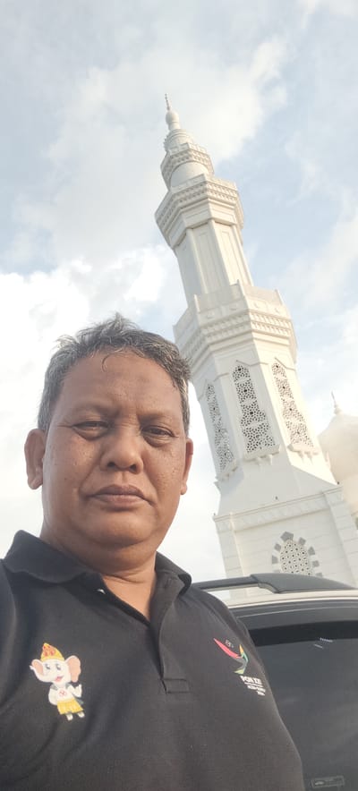 A close-up, low-angle, outdoor selfie features a middle-aged East Asian man in the foreground, wearing a black polo shirt with a cartoon elephant emblem on the left chest and a logo with text on the right. His face is looking directly at the camera, with a neutral expression. His hair is graying and tousled, and his skin appears weathered with some texture. Behind him, a tall, white minaret of a mosque dominates the background, reaching towards a cloudy sky. The architecture of the minaret is intricate, with decorative patterns and a pointed spire. The setting appears to be the city of Ranto, Indonesia, given the presence of the mosque. The lower portion of a vehicle is visible to the right of the man, partially obscuring the lower part of the minaret. The lighting suggests it is daytime, possibly late afternoon, with soft sunlight filtering through the clouds.