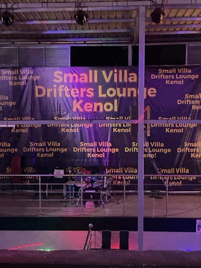 The image captures a nighttime scene at an outdoor or semi-outdoor venue, likely in Kimorori, Kenya, given the visible text and provided context. The central feature is a large purple banner with prominent yellow and orange text, repeatedly displaying "Small Villa Drifters Lounge Kenol." Metal scaffolding or railings are visible in the foreground and midground, partially obscuring the banner and creating a layered effect.
To the right, a stage area is set up with musical instruments, including a drum set featuring a clear "YAMAHA" logo. Red light beams or lasers cut across the stage and a distant wall, contributing to the evening ambiance. Wall-mounted lights are also visible on the left side of the frame. The overall impression is that of a venue prepared for a live music event or a promotional setup for a lounge or club. No people are discernible in the frame. The artificial lighting against the dark background confirms it is nighttime.