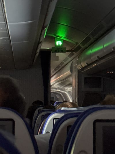 The image depicts the interior of an Aerobus shuttle at Sheremetyevo International Airport in Stolbishche, Russia. The perspective is from inside the carriage, looking lengthwise through the vehicle. In the foreground, the corner of a seat back is visible, with a blue and white frame. To the left, the interior walls of the shuttle are visible with fluorescent green lighting strips. A sign is visible, presumably in Russian, possibly indicating an exit. Further down the length of the shuttle, a series of seats and windows are discernible, diminishing into the distance.