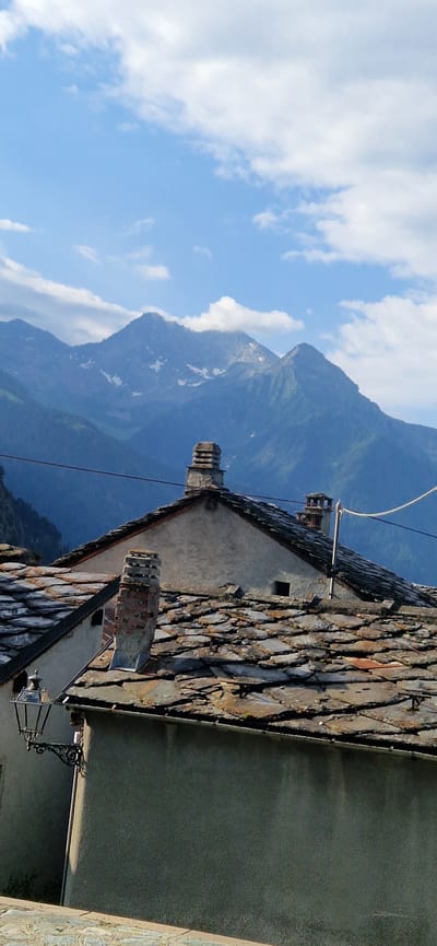 The image captures a view of rooftops and mountains in Brusson, Italy. In the foreground, a grey stucco building with a stone-tiled roof is visible. A vintage black lantern with intricate scrollwork is mounted on the left side of the building. A stone chimney with a weathered, layered cap stands on the roof. Further back, another building with a similar stone-tiled roof and two chimneys is present. The background is dominated by majestic blue mountains, some with patches of snow, under a bright sky with scattered white clouds. The overall scene conveys a sense of rustic charm and natural beauty characteristic of a mountain village.