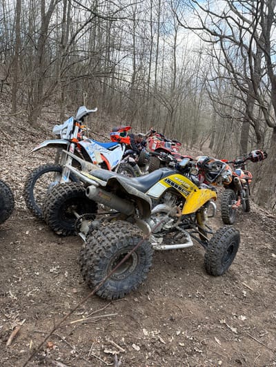 The scene depicts a group of off-road vehicles parked on a dirt trail within a dense, leafless deciduous forest near Kozichino, Bulgaria. In the foreground, a yellow Yamaha Raptor ATV, covered in mud, is prominently positioned with its rear facing the viewer. Its knobby tires and exhaust system are clearly visible. To its immediate left, a white and orange KTM dirt bike is parked, with an orange, black, and white helmet resting on its seat. Further into the midground, behind the yellow ATV, a red ATV is partially visible, also appearing muddy. Another helmet is situated near its handlebars. In the background, to the right, at least two more off-road vehicles (one appearing orange/red) are partially discernible. Two individuals, wearing dark riding gear, are faintly visible between the white KTM dirt bike and the red ATV, standing near the vehicles. The forest floor is composed of damp earth, scattered with fallen leaves and twigs. The trees are bare, indicating late autumn, winter, or early spring. The sky overhead is partly cloudy. The terrain is an unpaved, sloping path, suggesting a designated off-road riding area.