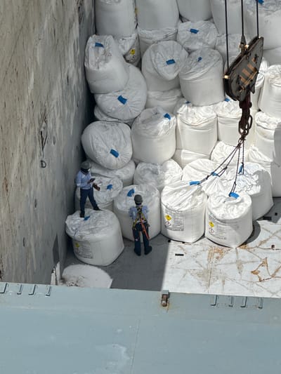 In Uribia, Colombia, inside the hold of a cargo ship, two individuals in safety gear are overseeing a large shipment of white bulk bags, likely containing industrial materials or agricultural products. One individual, wearing a blue shirt and dark pants with a hard hat, stands on top of a bag and appears to be using a tablet. The other individual, in a full safety harness and helmet, is positioned on the deck, facing away from the camera. The bags are stacked high, filling the space, with a crane mechanism visible at the top right of the frame, suggesting the ongoing loading or unloading process. The environment is industrial, with the ship's hull and deck forming the background. The lighting suggests it is daytime.