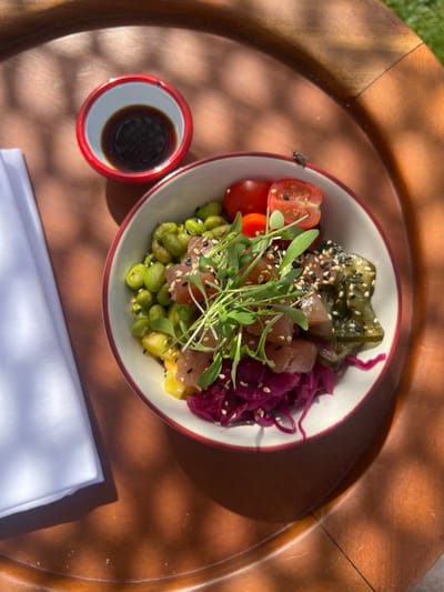 A high-angle, overhead shot depicts a poke bowl and accompanying items on a light brown wooden surface, likely a table or tray, located outdoors in Aljezur, Portugal, under direct sunlight, evidenced by distinct shadows.

The central subject is a white bowl with a thin reddish-brown rim, filled with various ingredients. Visible components include diced raw tuna, shelled green edamame beans, halved red cherry tomatoes, shredded dark red pickled cabbage, yellow fruit chunks (possibly pineapple or mango), and a garnish of green microgreens. The entire dish is topped with a sprinkling of white and black sesame seeds.

To the upper left of the bowl, a small, round white ceramic dish with a prominent red rim contains a dark liquid, consistent with soy sauce. A folded white cloth napkin is positioned to the left of the bowl and the sauce dish.

A single housefly (Musca domestica) is perched on the outer rim of the poke bowl, positioned above the cherry tomatoes. The wooden surface displays a visible grain and reflects sunlight, creating areas of bright highlights and deep shadows. No individuals are present in the frame.