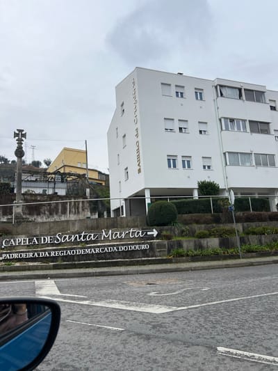 The image displays an urban street scene in Santa Marta de Penaguião, Portugal, under an overcast sky. In the immediate foreground, the lower left quadrant reveals the clean side mirror of a blue vehicle, reflecting a blurred interior. The road surface is gray asphalt with white painted directional arrows and lines. A multi-level concrete sign structure occupies the mid-ground, prominently displaying "CAPELA DE Santa Marta" with a right-pointing arrow on its upper tier, and "PADROEIRA DA REGIAO DEMARCADA DO DOURO" on its lower tier. This sign is set into tiered landscaped planters containing manicured green bushes. To the right of the sign, a modern, five-story white building with "BAIRRO DE CRISTO" written vertically in gold lettering on its left facade is visible. The building features multiple rectangular windows, many equipped with closed or partially closed white external roller shutters. To the left of the sign, on a raised, terraced area, a stone cruzeiro (cross monument) with an ornate, double-barred cross finial is present. Behind the cruzeiro, a yellow two-story building with visible metal scaffolding or trellises is partially obscured on higher ground. Utility poles and power lines traverse the background hillside. The sky is uniformly gray and cloudy. No individuals are visible within the scene.