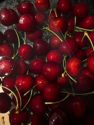 The image is a close-up, overhead shot of a pile of fresh, dark red cherries submerged in water. The cherries are plump and glossy, with some showing water droplets on their surfaces. Several green stems are visible, some detached and others still attached to the cherries. The background appears to be a dark, reflective surface, possibly a bowl or sink. The lighting is somewhat dim, creating highlights on the cherries and obscuring some details in the shadows. There are no people, animals, or specific landmarks visible that would indicate a location in Amstelveen, Netherlands. The image focuses solely on the texture and color of the cherries.