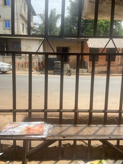 The image presents a street view from an interior vantage point, looking out through dark metal bars, likely a window grille or gate, in Ojoto, Nigeria. In the foreground, a worn wooden bench occupies the lower section of the frame, with multiple plastic-wrapped items, appearing to be colorful magazines or posters, resting on its left side.
Beyond the bars, a paved road extends horizontally across the scene. A silver four-door sedan is partially visible, parked on the left side of the road. Across the street, a line of multicolored fabric strips or ribbons is suspended against a light-colored building or wall. To the right of these strips, a large black plastic water tank stands next to a stack of four used vehicle tires. Adjacent to the tires, a small, dark-colored, two-wheeled trolley or cart is positioned on the ground.
The background features several buildings and natural elements. On the far left, a multi-story structure with light-colored walls and blue-tinted windows is visible. In the center-right, two single-story, earth-toned buildings are present. The left-most of these has an open doorway accessible via a rough, stone or concrete ramp/steps. The building to its right features a corrugated metal roof and what appear to be closed window openings. Various green trees, including a tall palm tree, are situated behind and between these structures, under a bright, largely overcast sky. No individuals are visible or engaged in activity within the frame.