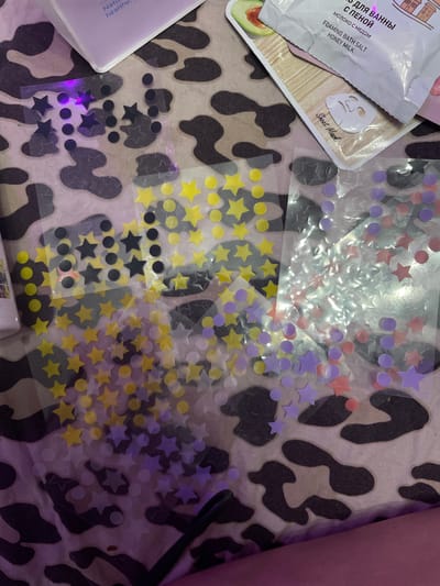 The image depicts a close-up, overhead view of several sheets of star and circle stickers scattered across a textured, possibly fabric, surface with a cow-print pattern. The stickers are in various colors including yellow, black, purple, and pink. Some of the stickers appear to be transparent or holographic. In the upper portion of the image, a portion of a white and light purple container or packaging is visible, with text that is partially obscured and in a foreign language. There is also a corner of a face mask packet visible, also with foreign text. The lighting suggests an indoor setting, with a noticeable purple hue casting over parts of the scene, possibly from artificial lighting. The image does not provide any information or context about the city of Romanovka, Russia.