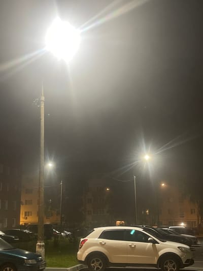 An outdoor nighttime photograph captures a street scene in Shakhovskaya, Russia, characterized by significant atmospheric haze and the illumination from streetlights. In the foreground, several parked cars are visible, including a white SUV and a dark-colored sedan. The backdrop consists of multi-story residential buildings, their windows dimly lit. A prominent wooden utility pole with attached wires stands to the left of the frame, illuminated by a bright streetlight at its top, which creates a lens flare effect. Another streetlight is visible further down the street. The overall environment appears to be a residential area, with a sidewalk and grassy patches. There are no people visible in the image.