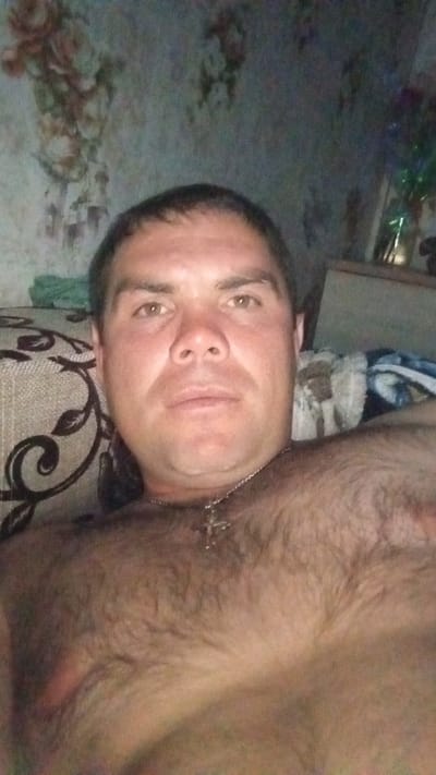 An overhead, close-up selfie shows a man from the chest up. He has short, dark hair, light brown eyes, and is wearing a silver chain with a cross pendant. The man is resting on a sofa with a patterned upholstery. In the background, there is a wall with a floral pattern wallpaper and a shelf with a vase of flowers. The image is grainy and appears to be taken indoors at night, possibly in Kochyovo, Russia.