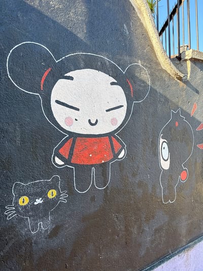 This image captures a portion of a dark grey, textured outdoor wall in Tomé, Chile, featuring a mural with three cartoon characters. Centrally positioned is Pucca, depicted with a large white face, smiling expression, and rosy cheeks, outlined in white with black "hair buns" accented by red crescents, wearing a red top with white outlines. To her lower left is a black cat with prominent yellow eyes, white whiskers, and a white outline, facing forward. To Pucca's right, the character Garu is shown in profile, facing left towards Pucca; he is black with a white eye, small red facial features, and red spikes emanating from his head. The bottom edge of the wall reveals a narrow purple baseboard. The top right segment of the image includes a light-colored concrete structure with a dark metal fence or railing and a small green plant, all partially illuminated by direct sunlight, which casts distinct shadows. The wall shows minor imperfections and variations in color saturation.