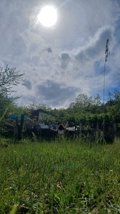 A medium, outdoor, wide shot oriented to the right depicts a sunny day in Gorna Verenitsa, Bulgaria. The foreground is dominated by tall, green grass that appears to be a field. In the middle ground, a house or building is partially obscured by trees, and someone is stretching. The sky in the background is filled with clouds, and the sun is brightly shining near the left side of the frame.