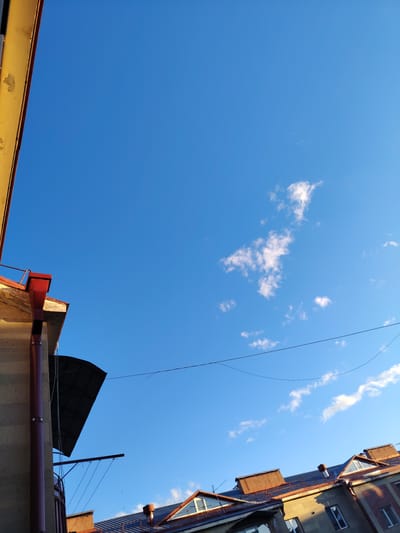 The image depicts a view looking up towards a clear blue sky from the street level in Border Gyumri-Akhuryan, Armenia. The top left portion of the frame is dominated by a weathered, yellow-painted structure with a red gutter system. Below this, the rooftops of several buildings are visible, characterized by reddish-brown tiled roofs and attic windows. Some of these rooftops also feature chimneys. Thin wires are strung across the sky in the middle and upper right portions of the image. A few wispy white clouds are scattered across the expanse of the blue sky. The sunlight suggests it is daytime, casting shadows on the buildings.