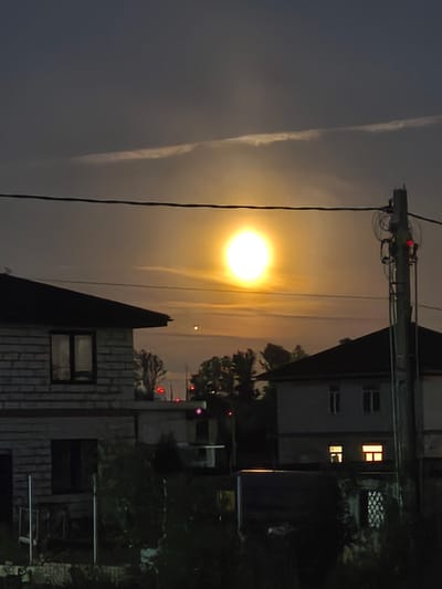 The image captures a nocturnal scene in Frontoviki, Russia, under a large, bright moon that dominates the sky. The moon's luminescence casts a warm, yellow-orange glow across the landscape, illuminating a strip of clouds that stretches horizontally. In the foreground, two residential buildings are visible. The building on the left is constructed with light-colored brick and features dark-framed windows. The building on the right, partially obscured by a utility pole, is a lighter color and also has dark-framed windows, some of which are lit from within. A utility pole with multiple wires and a red light fixture stands prominently in the right portion of the frame. Trees and dense foliage are visible between and in front of the houses. The overall impression is one of a quiet, dimly lit residential area at night.