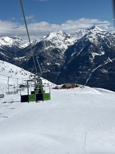 The image depicts a ski lift in Puy-Saint-André, France. The ski lift is in the foreground, with chairs featuring green and black upholstery. A steel cable runs along the upper left of the frame. The background showcases a snow-covered mountain landscape with a mix of forested and bare areas. The sky is blue with some white clouds. A building with a terrace, likely a mountain restaurant or station, is visible on the slope further up the mountain. Ski tracks are visible in the snow.