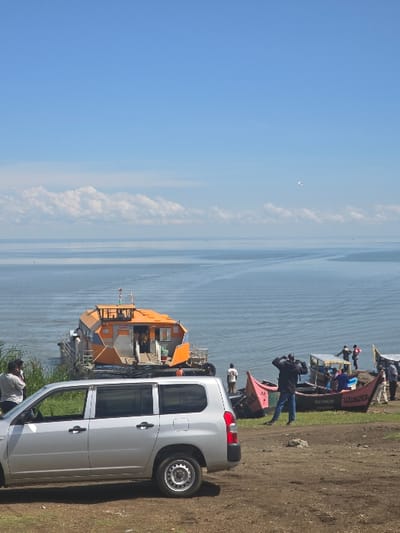 The image shows a scene from the shore of Lake Victoria in Homa Bay, Kenya, at the coordinates -0.522 latitude and 34.459 longitude. A silver Toyota Land Cruiser is parked on the shore, facing away from the lake. There is a small orange and yellow ferry docked near the shore. Several people are standing around the boat, and some are sitting in other smaller boats nearby. The sky is clear blue with some white clouds. The water is calm and there is green vegetation along the shore. The land is mostly brown dirt. The day appears to be sunny, with a light breeze.