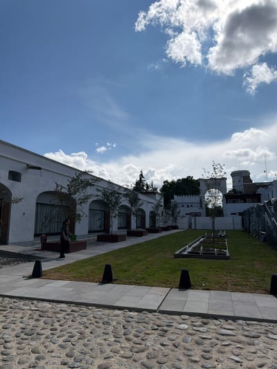 This image captures an outdoor scene in what appears to be a public or private courtyard, possibly in San Lucas el Grande, Mexico, given the architectural style. The scene is dominated by a clear blue sky with numerous fluffy white clouds.
On the left side of the frame, several white buildings with arched windows and dark frames are visible. Small, young trees are planted in rectangular planters along the base of these buildings, offering a touch of greenery against the white walls. One person is visible, sitting on a dark bench positioned in front of one of the buildings, appearing to relax. Further back, another white building features a prominent circular opening or archway, with a darker, possibly older structure behind it, partially covered by what looks like scaffolding or protective tarps. A small tower-like structure is also visible in the distance.
The right side of the image features a well-maintained green lawn, bordered by a light gray paved walkway made of square tiles. Adjacent to this walkway is a textured cobblestone path or area, composed of rounded, smooth stones. A long, rectangular planter box with small plants is embedded in the lawn further back. Several dark, triangular or pyramidal bollards are spaced along the edges of the paved areas.
The time of day appears to be daytime, with bright sunlight casting shadows, suggesting mid-morning or afternoon. The overall atmosphere is peaceful and serene. There is no visible text in the image.