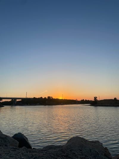A serene sunset over the waters near Nea Potidea, Greece, captures the tranquil beauty of the coastline. The sky transitions from a clear, deep blue at the top to a soft orange and yellow hue near the horizon, where the sun is setting. A prominent bridge, likely part of the Nea Potidea canal, stretches across the scene from the left, its structure silhouetted against the vibrant sky.

Along the distant shoreline, a line of trees and low-lying buildings creates a dark, undulating silhouette. Several vehicles and a sailboat are visible docked near the bridge, suggesting a marina or harbor area. The water in the foreground is calm, reflecting the warm colors of the sunset in gentle ripples. Large, rough rocks are scattered along the very edge of the foreground, creating a natural barrier between the land and the water. The overall atmosphere is peaceful and picturesque, with no people or animals visible in the frame.