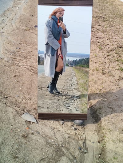 The image depicts a mirror standing on a dirt and gravel surface, reflecting a woman taking a selfie. The woman is wearing a grey overcoat, an orange dress, a blue scarf, and black boots. The background visible in the mirror reveals a landscape with trees, a body of water, and a building with a green roof in the distance. The surrounding environment consists of dirt, gravel, some sparse vegetation, and shadows from the sunlight. Based on the distant building and landscape, the location is likely Нечкино, Russia.