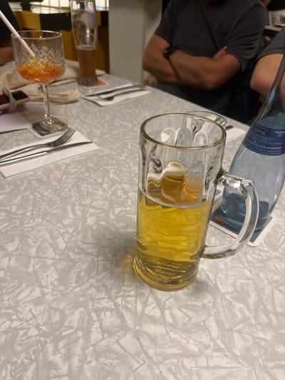 The image depicts an indoor dining or drinking setting, likely in a restaurant or pub in Tuttlingen, Germany, as indicated by "511 Tuttlingen" visible on a water bottle label.

In the foreground, slightly right of center, a clear glass beer mug, approximately half-full with a golden-yellow liquid and a minimal head, rests on a white square coaster. To its right and slightly behind it, a transparent blue plastic bottle, labeled "MINERALWASSER" and "511 Tuttlingen," also sits on a white square coaster.

In the mid-ground, to the left, a stemmed glass with a faceted design holds an orange-colored beverage containing ice cubes and a white straw. This glass is placed on a round green and white coaster. Adjacent to this glass, to the left, a small portion of a smartphone is visible resting on a white napkin. Further left, a silver fork and knife are neatly arranged on a folded white napkin.

In the background, on the right side, a person's torso and arms are visible. The individual wears a dark grey short-sleeved t-shirt and a dark wristwatch on their left wrist, with their arms crossed. Behind the stemmed glass on the left, another tall, slender glass containing a brown liquid is partially visible. The background also shows indistinct yellow and dark architectural elements. The table is covered with a light-colored, patterned tablecloth.