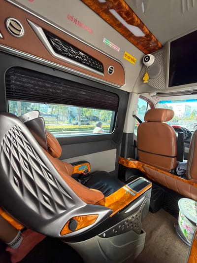 This is a media file taken inside a passenger van, possibly a sleeper bus, with a focus on the interior furnishings and a view through the window. The van appears to be modern and luxurious, with plush brown leather seats that recline, offering a comfortable travel experience. The interior is decorated with wood-grain accents and has a sleek, contemporary design.
Visible objects include individual passenger seats, a large screen at the front, a small camera mounted on the wall, and storage compartments. There are also some decals with Vietnamese text, one of which reads "TÍNH MẠNG CON NGƯỜI LÀ QUAN TRỌNG NHẤT," meaning "Human life is the most important." Another decal says "XE HỢP ĐỒNG," indicating a contract vehicle.
Through the window, a daytime scene is visible, suggesting the van is in motion. The view shows trees, buildings, and a street, indicating an urban or suburban environment. The lighting and the clear sky suggest it is daytime, and the weather appears to be pleasant.
The overall impression is one of comfort and safety, with the text reinforcing the importance of passenger well-being. The van's interior design suggests a premium service, likely for long-distance travel. The scene is static, captured from a fixed perspective within the vehicle, focusing on the passenger experience and the exterior world passing by.