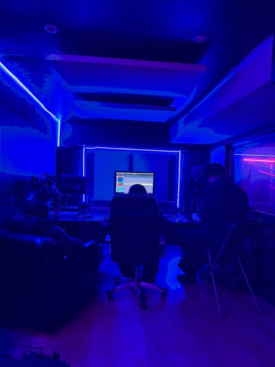 A dimly lit room bathed in blue light features soundproofing panels on the wall. A monitor displays vertical lines. Neon lights outline the room's edges and the frame around the monitor. An individual wearing a VR headset sits in a chair. The perspective is tilted.