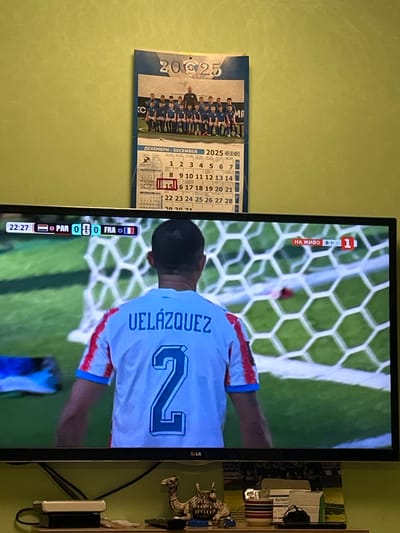 An interior, eye-level, medium shot captures a television screen displaying a soccer match, with a portion of a wall calendar above it. The television displays the score: PAR 0-0 FRA, with a game clock showing 22:27. The match is live, indicated by "НА ЖИВО" and "1". A soccer player wearing a white jersey with red and blue trim and the name "VELÁZQUEZ" and the number "2" on his back is visible from behind. Above the television, a wall calendar for December 2025 is partially visible, featuring a photograph of a youth soccer team. The wall behind the television is painted a light green. Below the television, on a shelf, are various electronic devices and decorative items, including a camel figurine. The location is Каменар, Bulgaria.