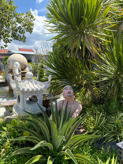 This is a medium shot photograph taken outdoors on a sunny day in Tuy An Nam, Vietnam. The scene features a statue of a bald, bearded man with his eyes closed, appearing to be in a meditative pose. He is partially obscured by lush green foliage, including large palm-like leaves and smaller succulents. Behind the statue, a stone pagoda-style lantern sits next to a tiered fountain topped with a large spherical object and a dragon sculpture. The background includes residential buildings with red-tiled roofs and a clear blue sky with scattered clouds. The overall setting is a garden or courtyard.