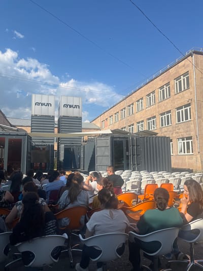 This media file shows an outdoor gathering of people sitting on chairs, possibly attending an event or presentation. The scene is set against a backdrop of a multi-story building with many windows and several shipping containers that appear to be used for the event space. The sky is blue with some clouds, suggesting it is daytime and the weather is fair.

The people are seated in rows of modern chairs, some of which are orange and white. Most of the individuals are facing away from the camera, with their backs turned as they look towards a central area, implying an ongoing activity. The composition suggests a sense of community or shared experience.

Two prominent signs are visible on the shipping containers, displaying the word "Yout" and "Anman" vertically, along with some Cyrillic or Armenian text that is partially legible. The overall impression is one of an organized event in an urban or semi-industrial setting in Gladzor, Armenia. The sunlight indicates it is likely afternoon, and the mood seems neutral, focused on the event at hand.