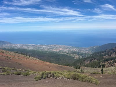 The image captures a panoramic view from a high vantage point, likely a volcanic slope, overlooking the coastal city of La Orotava, Spain. In the foreground, the terrain is a mixture of dark, volcanic soil and sparse, low-lying green shrubbery. The ground slopes downwards, showing patches of reddish-brown soil, characteristic of volcanic ash. Further down the slope, the vegetation becomes denser, transitioning into a dark green pine forest that covers the hillsides.

Beyond the forest, the city of La Orotava sprawls across the landscape, a densely packed collection of buildings with red-tiled roofs, interspersed with green areas. The city extends towards the coast, where it meets the vast expanse of the deep blue Atlantic Ocean. The ocean stretches to the horizon, meeting a bright, pale blue sky filled with wispy white clouds. The overall impression is one of a dramatic transition from rugged, arid volcanic terrain to a lush, populated coastal environment. There are no people or animals visible in the image.