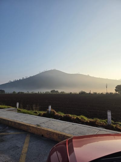 The image depicts a landscape in San Andrés Payuca, Mexico, featuring a mountain range partially obscured by haze under a blue sky. To the right of the frame, a plowed field with sporadic vegetation extends from the base of the mountain. A concrete curb borders the field, leading to a paved surface with yellow markings. A portion of a red vehicle is visible on the right edge of the frame, suggesting the viewpoint is from inside or adjacent to a parked car. Utility poles are placed intermittently along the roadside.