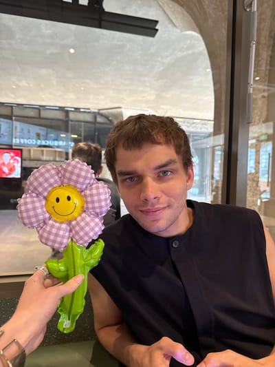 A man with blue eyes, wearing a dark shirt, holds a flower balloon with a yellow smiley face in front of him. The balloon flower has purple and white checkered petals and a green stem. An arm holds the balloon, with a ring on one finger and a watch on the wrist. The background shows a blurred interior of a cafe, with windows and possibly advertisements visible. This could be taken in Hongkou District, China.